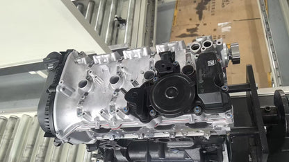 A silver and black automotive engine block assembly photographed indoors on a workbench or display stand, with metal shelving and cardboard boxes visible in the background.