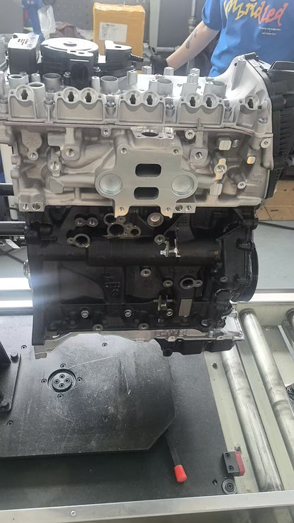 A large silver and black automotive Audi VW EA888 engine block mounted on a workbench in a workshop, with a person wearing a blue shirt partially visible in the background.