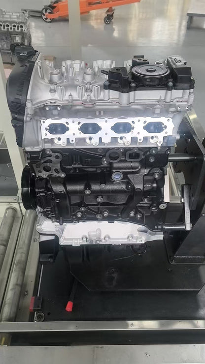 A large silver and black automotive engine block with visible cylinder head openings and mounting hardware, photographed on a metal support stand in an industrial workshop setting.