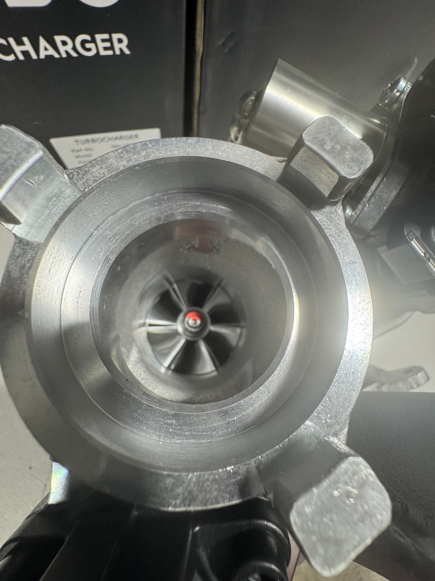 A close-up of a metallic turbocharger impeller wheel inside a silver metal housing, with a black box partially visible in the background.