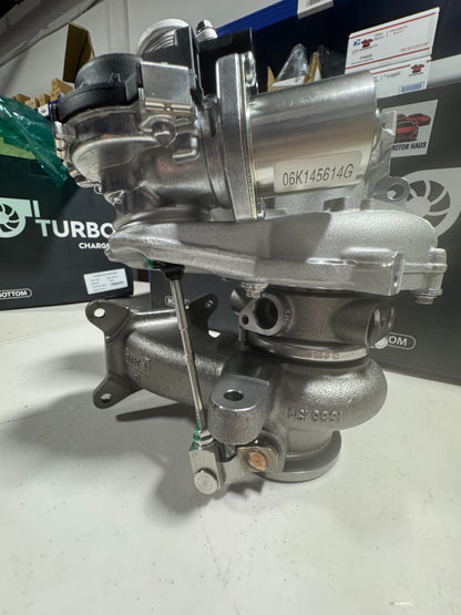 A new automotive turbocharger with a silver cast metal body, featuring a white sticker with part number '06K145614G' on the compressor housing, photographed on a white surface with a workshop background.
