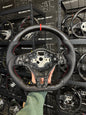 A carbon fiber racing steering wheel with red stitching and a black leather-wrapped rim, held by a hand in front of a wall of Scenestro branded steering wheels.