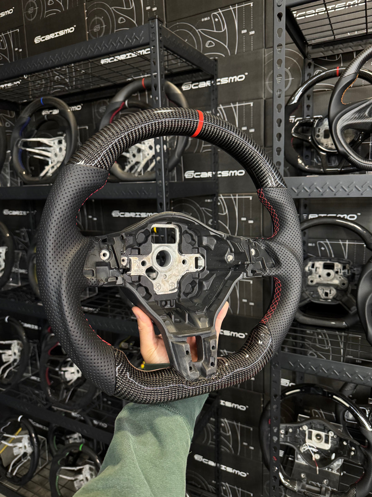 A carbon fiber racing steering wheel with a black leather wrap and red stitching, held by a hand in front of a wall of Scaramuzi branded display shelves.
